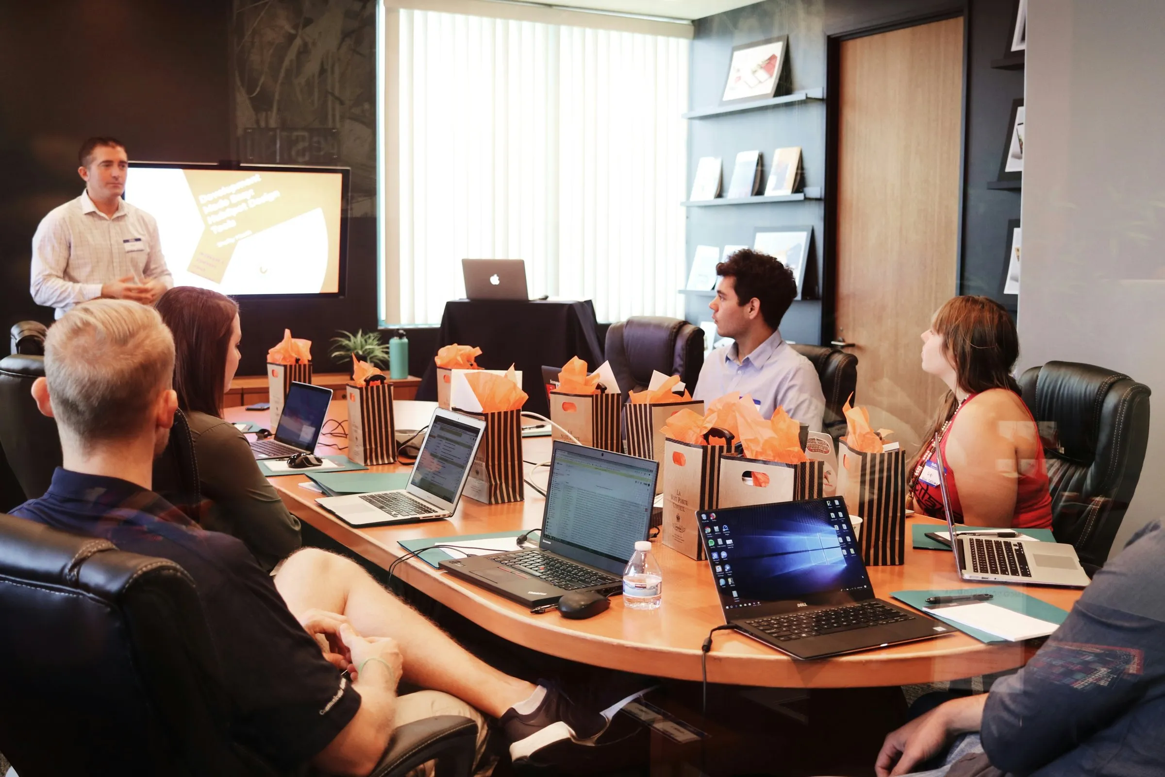 A presenter at a screen, briefing a meeting around a boardroom table