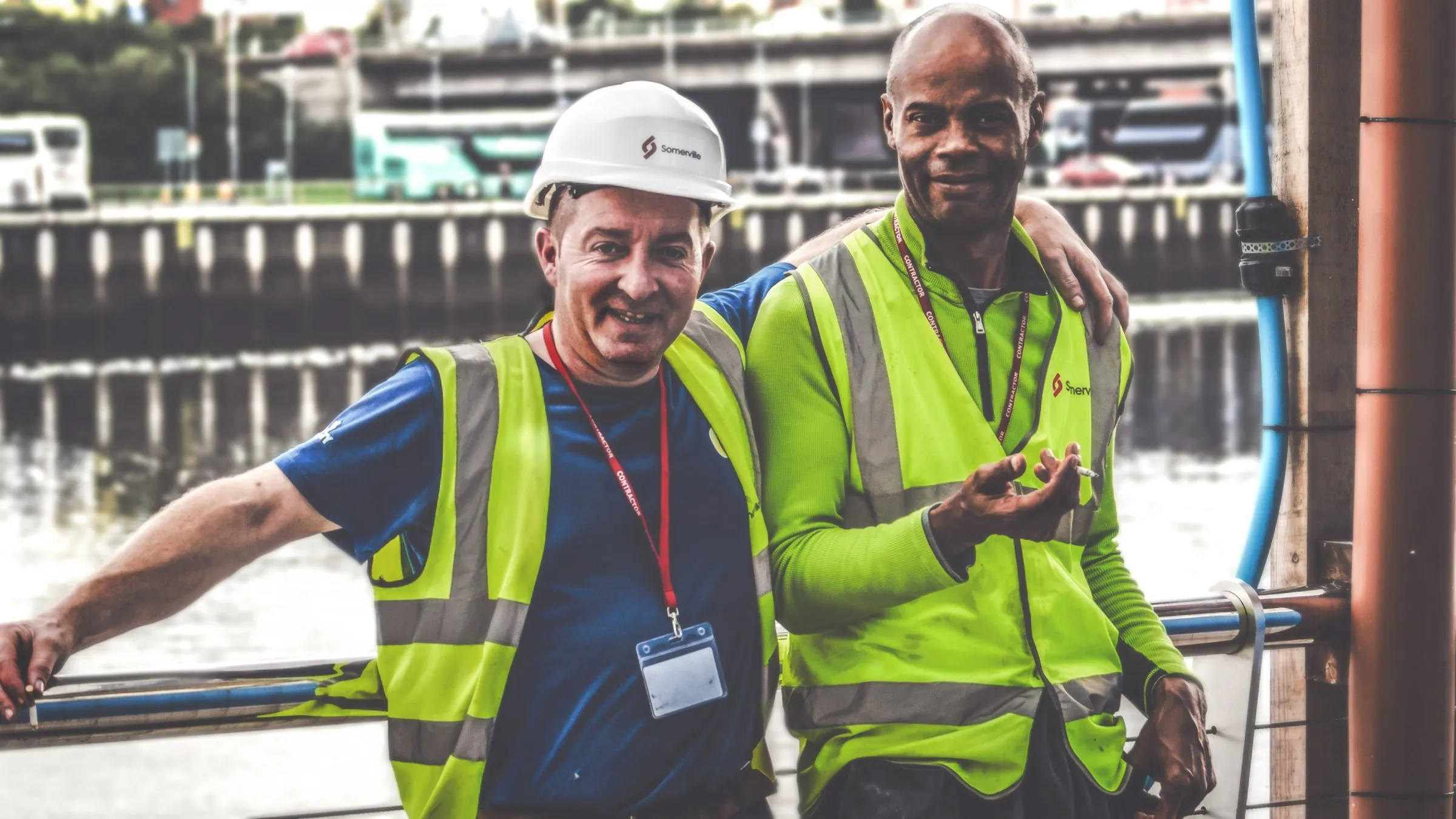 Two smiling workers in hard hats and high-visibility vests by a harbour
